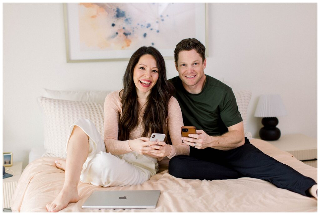 Arizona Brand Photographer couple smiles at the camera, they are sitting in bed holding cell phones.