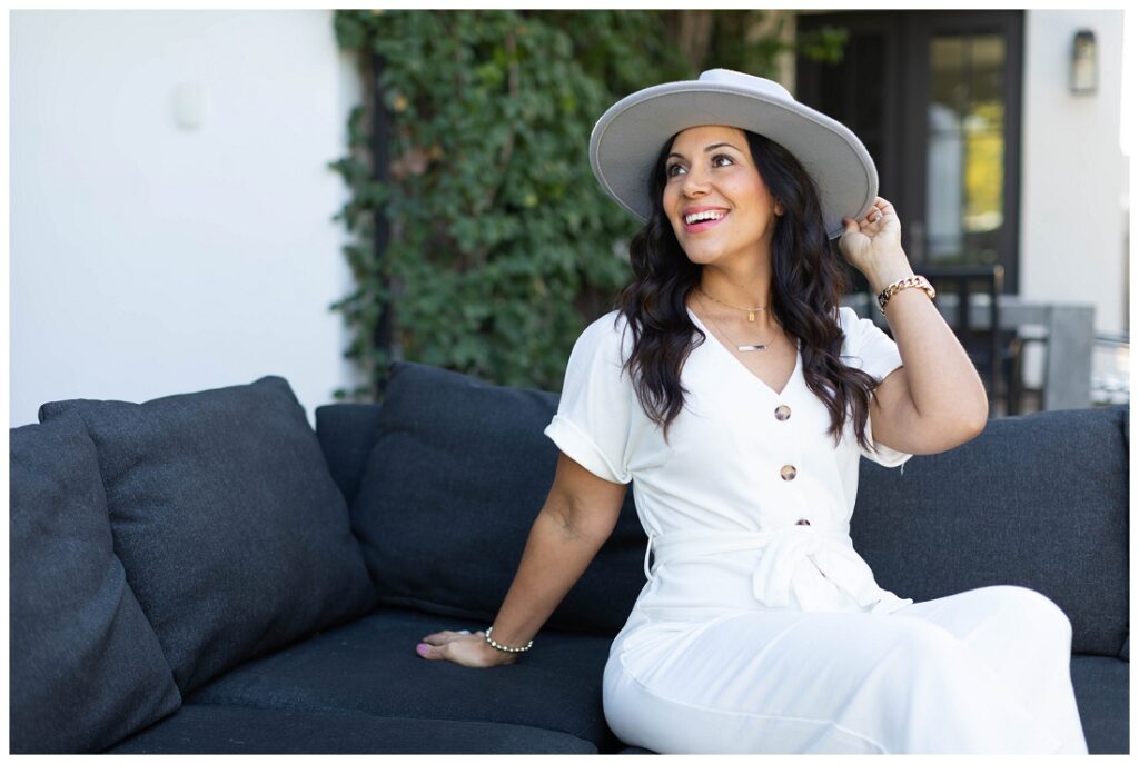 Denise Karis Brand Photography Arizona woman smiles at the sky, wearing a white hat and white dress while sitting on a navy sofa outdoors. 