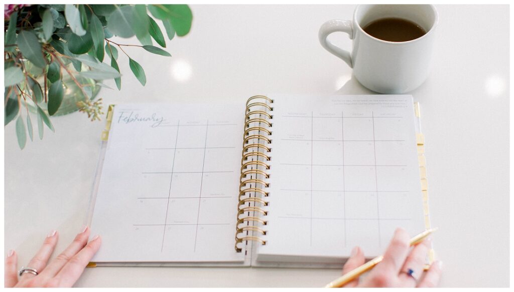 a planner lies open on a counter with a plant and a coffee mug in the upper corners, a hand is at the lower right holding a pencil Denise Karis brand photography 