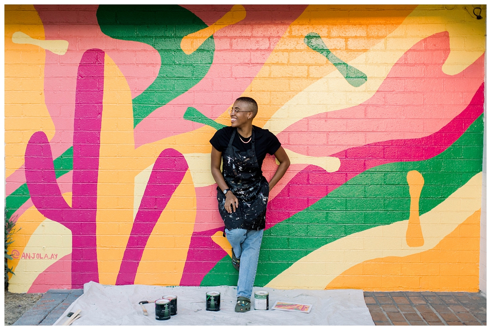 Woman in a painter's apron stands in front of a colorful mural
