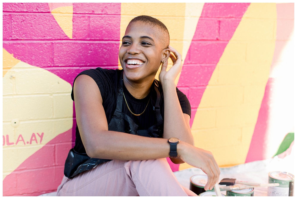 a woman in a black shirt and pink pants smiles off to the side of a camera. in the background is a large painted mural