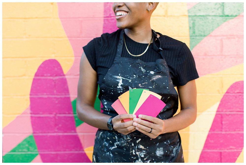 A painter in a painters apron holds colorful paint swatches and stands in front of a large colorful mural