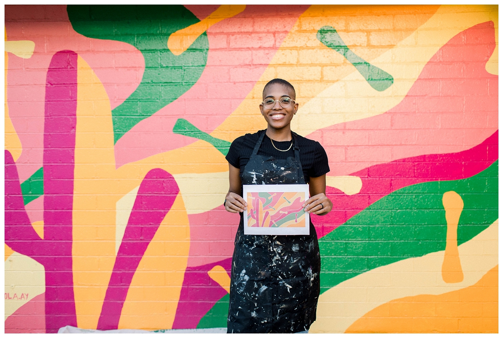 A painter in a painters apron stands in front of a large colorful mural and holds a printed, smaller version of the mural in her hands. 