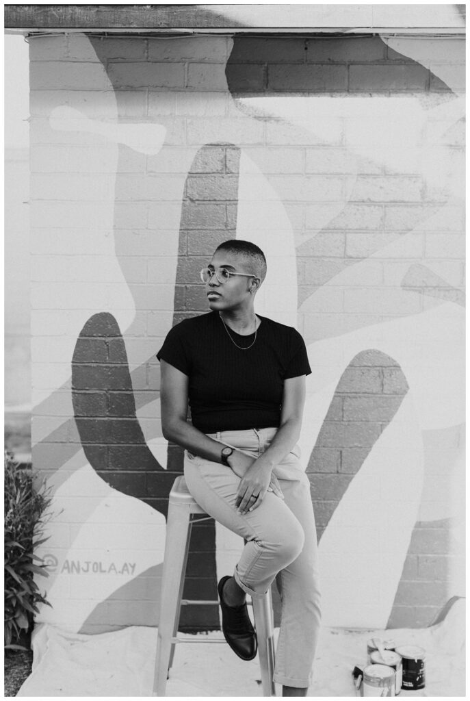 a black and white image of a painter in a black shirt and pants sits on a stool in front of a large mural. 