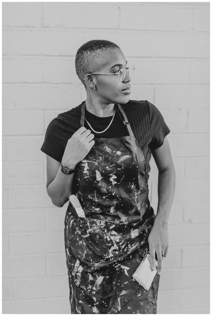 a black and white image of a painter in a painters apron stnading in front of a white brick wall. 