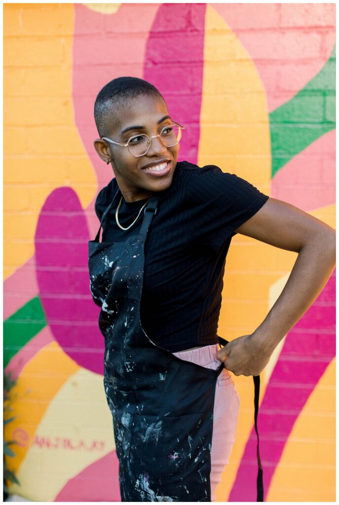 A painter ties an apron around her waits while standing in front of a large colorcul mural 