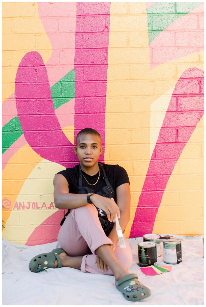 A painter in a black top and pink pants sits in front of a large colorful mural 