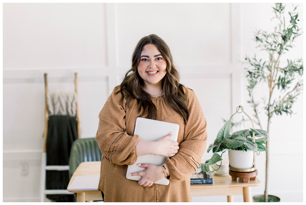 A woman in a mustard top holds a notebook and stands in front of a desk