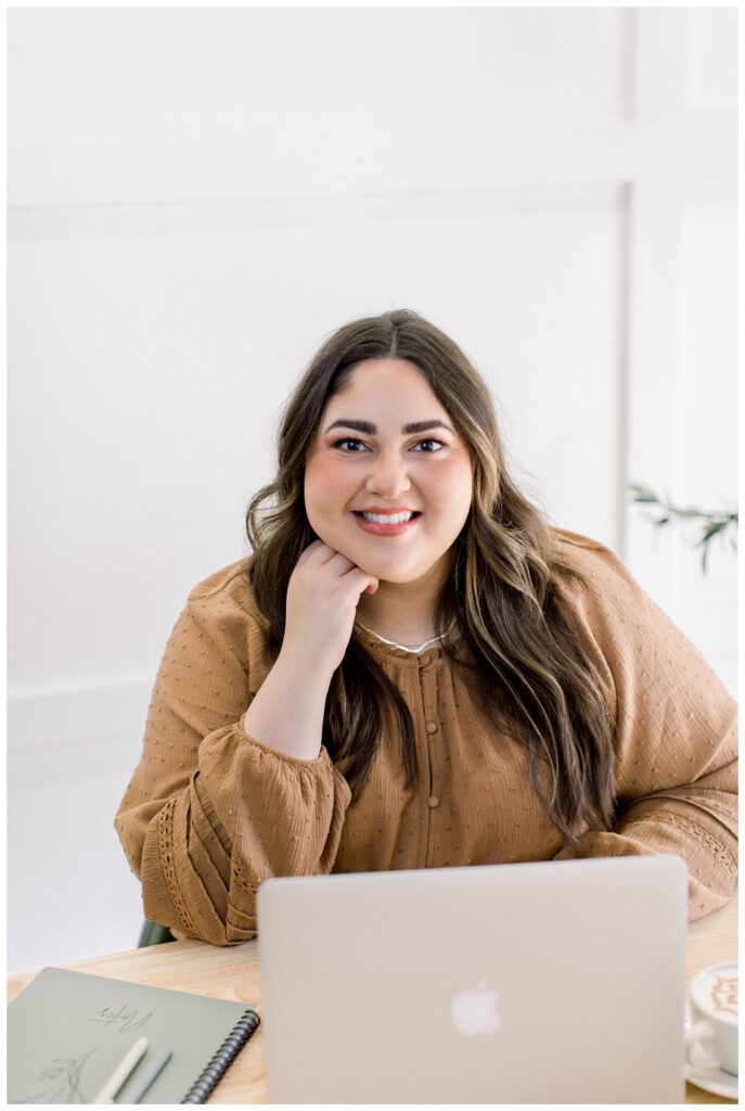 A woman in a mustard colored top has her hand on her chin and sits at a desk in front of an open laptop. She smiles at the camera. 
