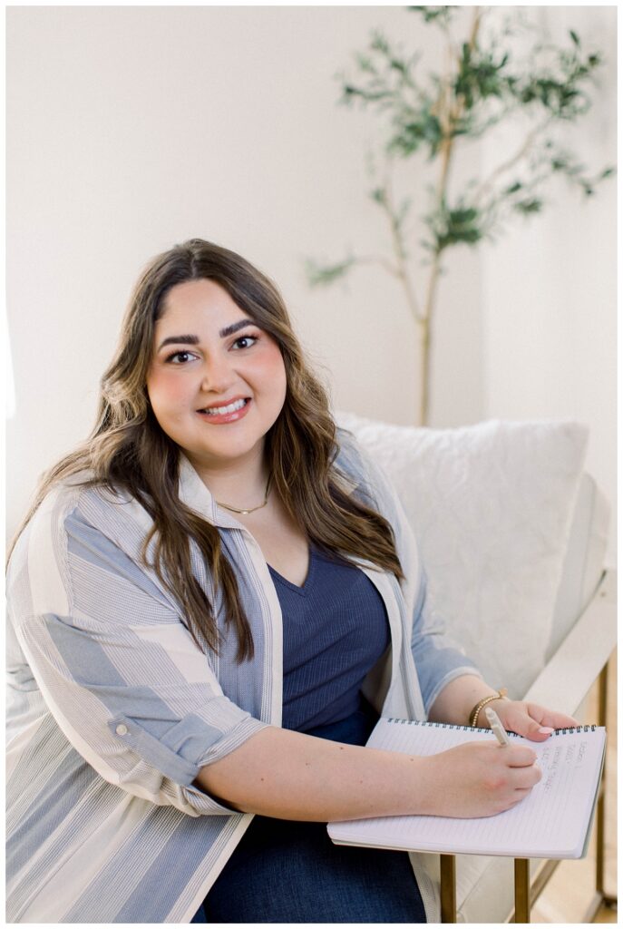 A woman in a blue top sits on a chair writing in a notebook. She is smiling at the camera. 