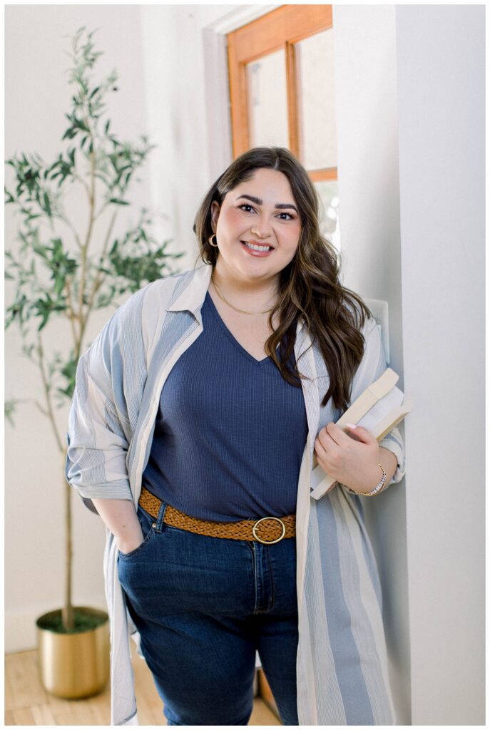 A woman in jeans and a blue top holds a notebook and leans against the wall of a studio office