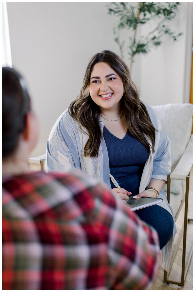 A womain wirting in a notebook sits on a chair and talks to a client. We only see the clients shoulder in the foreground of the image. 
