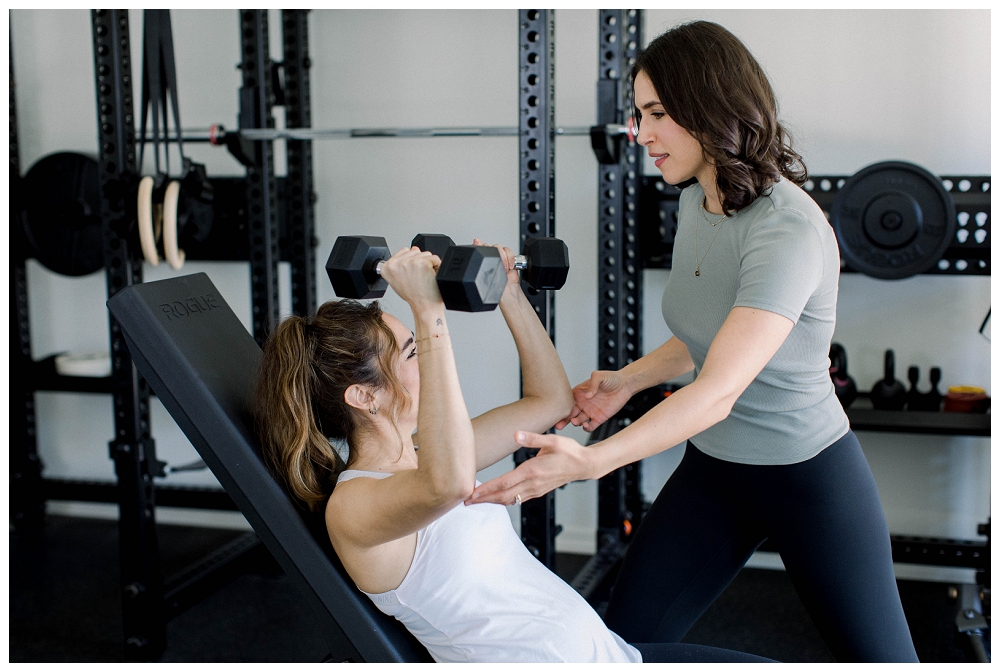 A woman helps another woman exercise in a gym setting. 