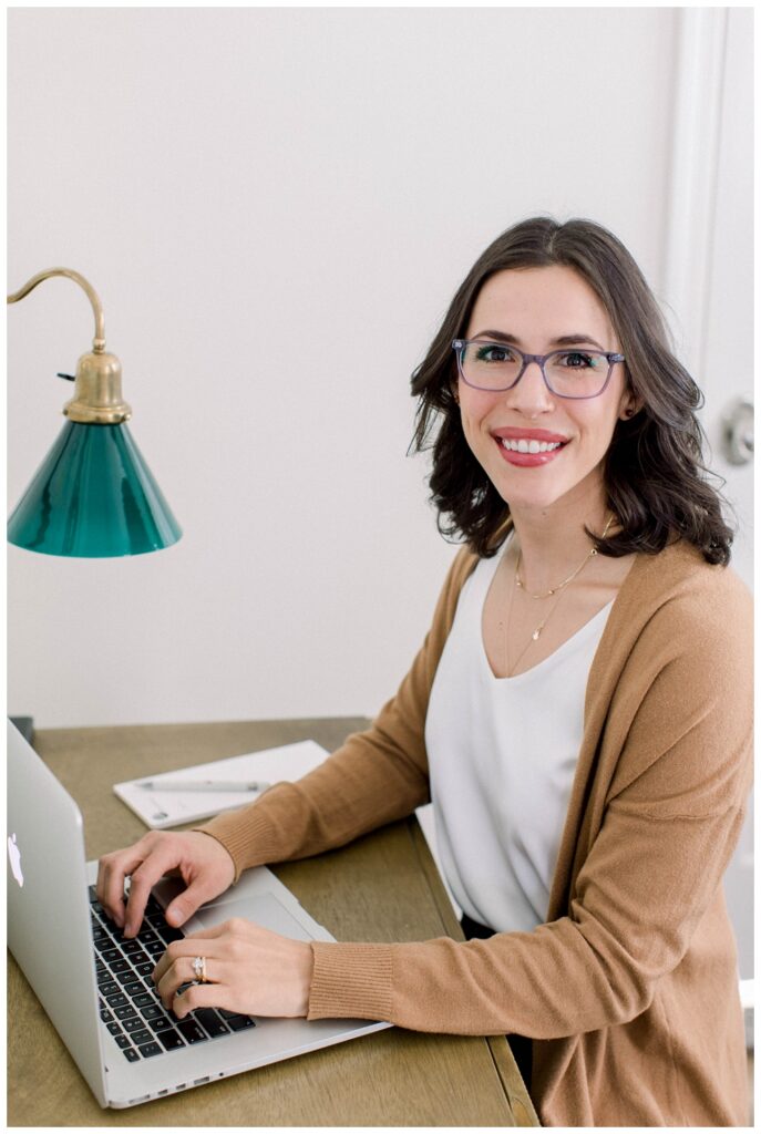 A woman wearing glasses and a beige sweater sits at a desk typing on a laptop. She smiles at the camera. 