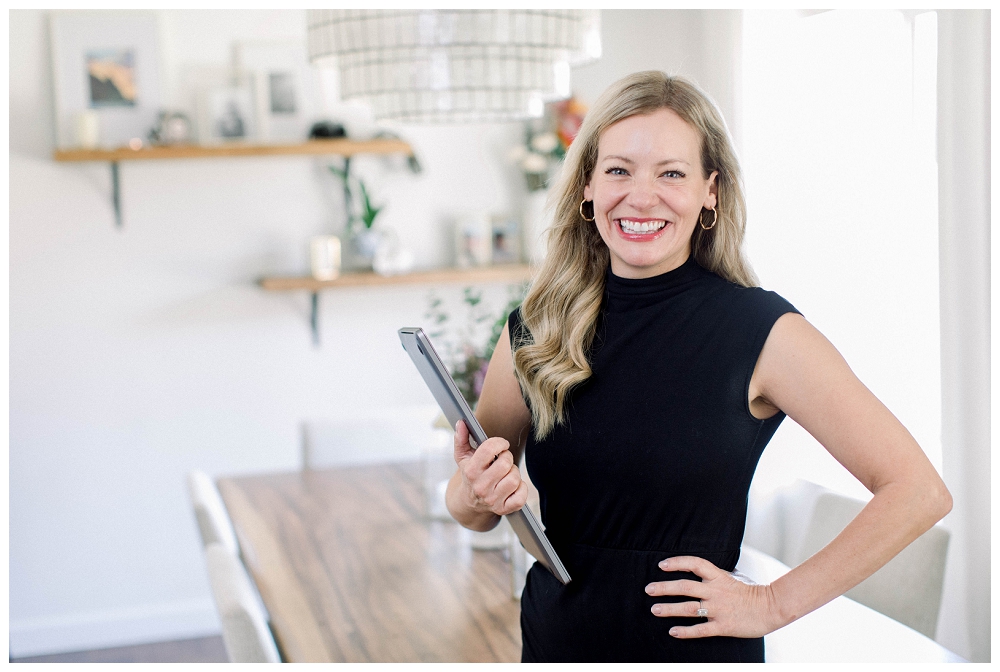 Branding portrait of a woman smiling beside a laptop at a wooden table in a bright home office.