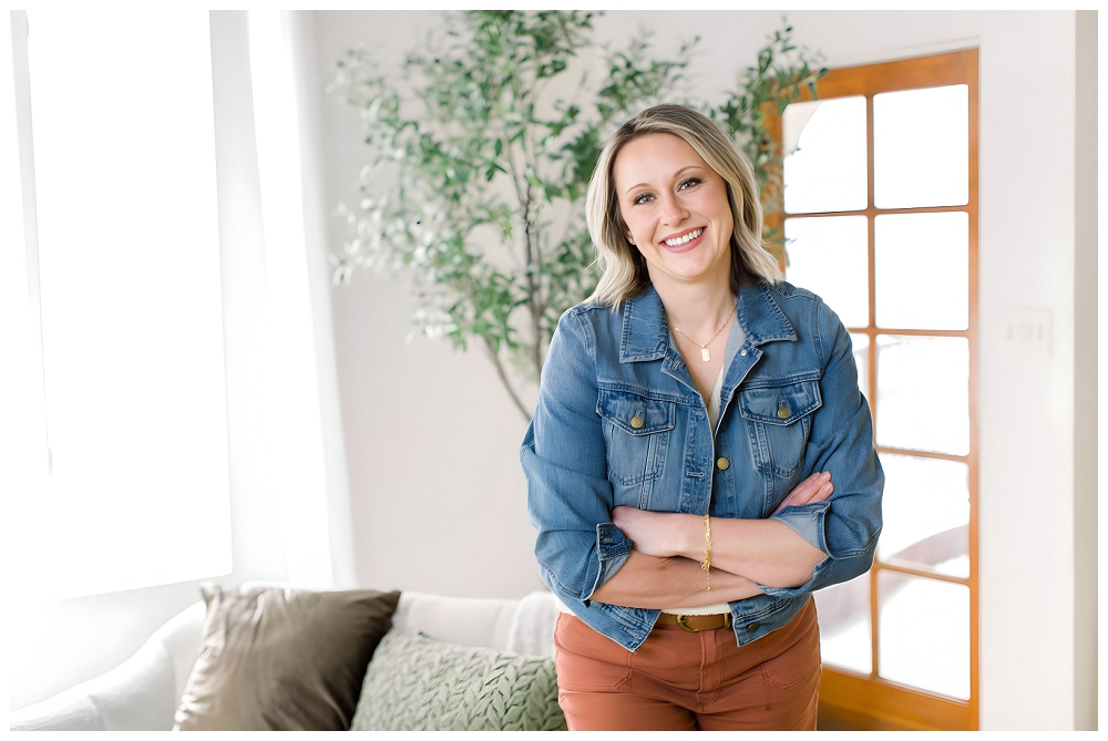 Woman wearing a denim jacket and orange pants smiles at the camera. There's a living room setting behind her and she has her arms folded. 