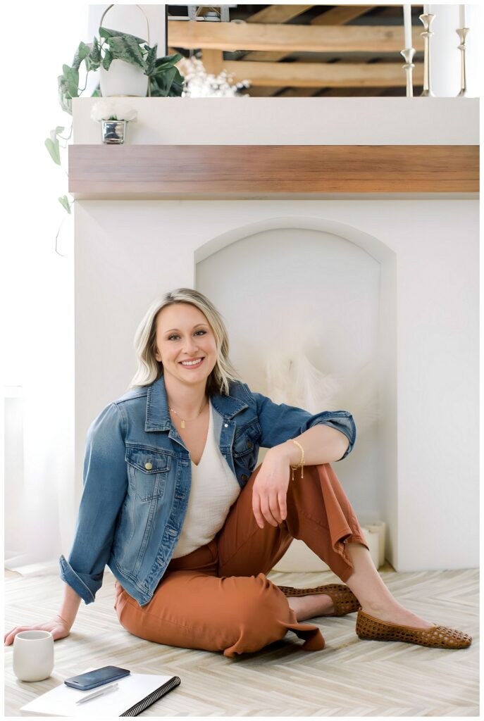 A woman in a denim jacket and orange pants sits in front of a fireplace and smiles at the camera. 