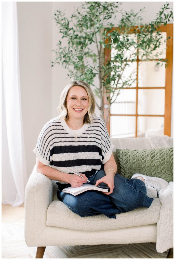 Brand photography, a woman in a white and navy stripped top and jeans sits on a sofa with a notebook open in her lap. Her hand is positioned to write. 