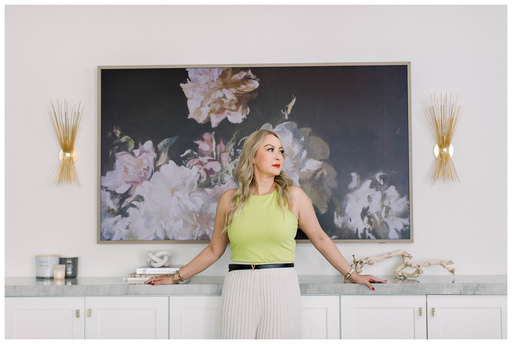 A woman in a bright lime top and white pants stands in front of a floral painting. 
