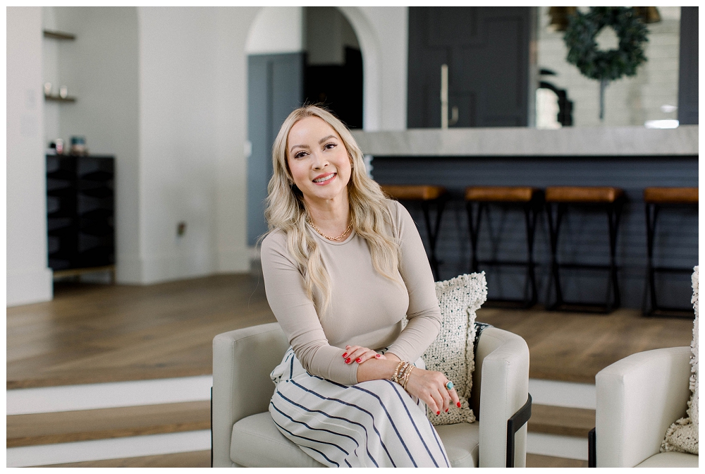 blond woman in a beige top and striped pants sits in a tan chair. there is a luxury bar in the background with navy paint and leather stools.