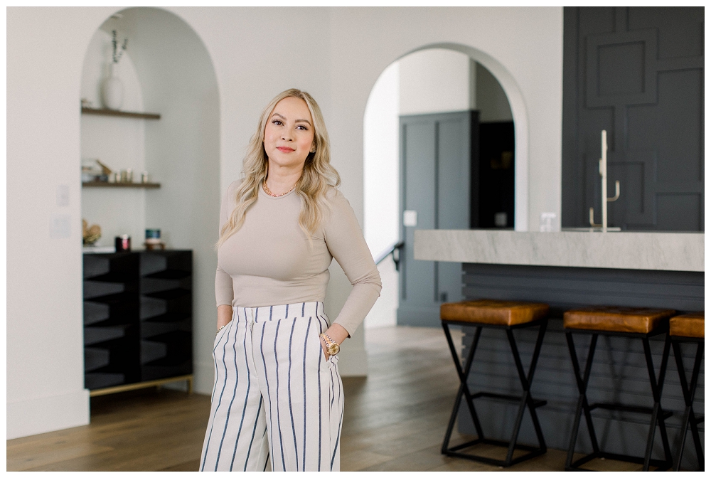 blond woman in a tan top and stripped pants stands with her hands in her pockets. There's a luxury bar in the background with leather stools and navy paint.  
