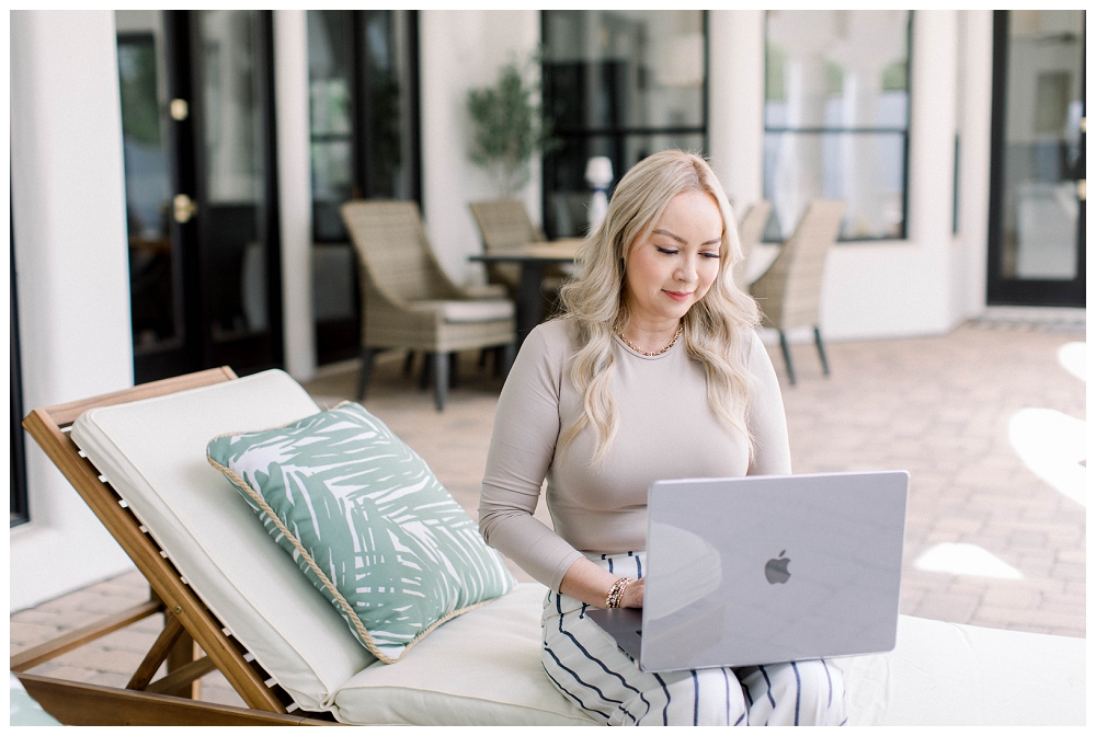 A woman sits outdoors on a recliner chair. She is working on a laptop and wears a beige top and stripped pants. 