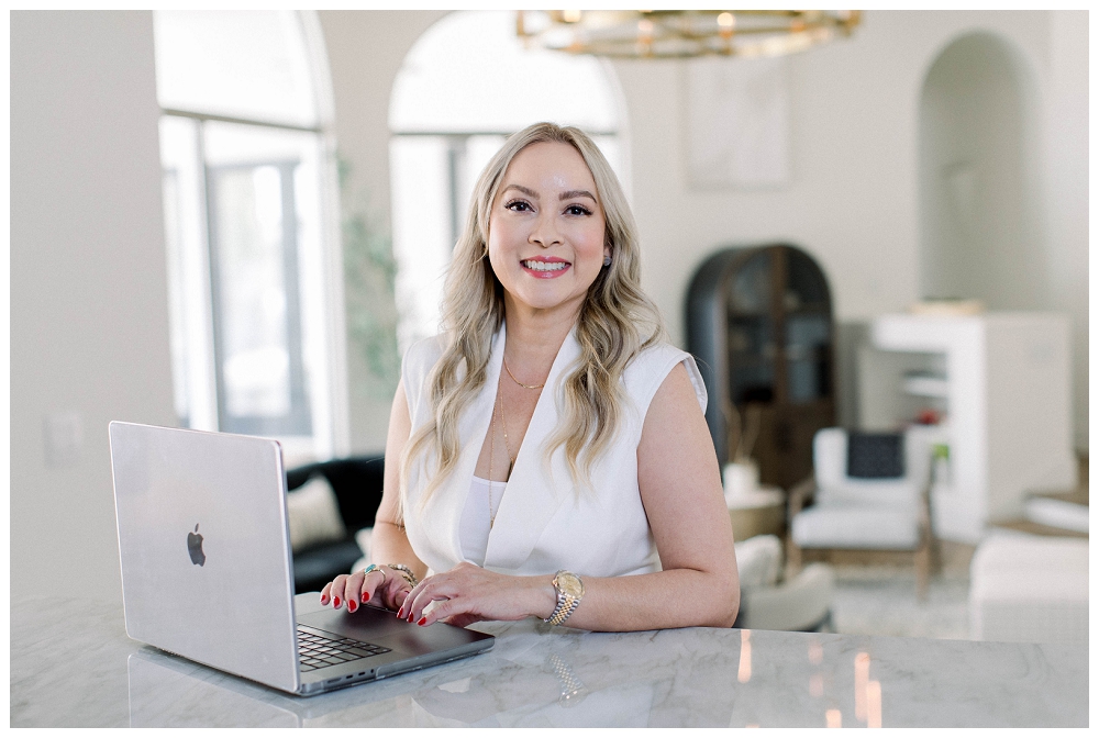 A woman sits at a table on a laptop smiling at the camera. There is a living room behind her. 