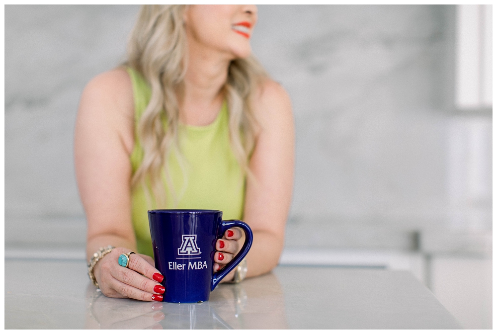 A woman leans on a counter. The photo is focused on a mug from a university in her hands. 