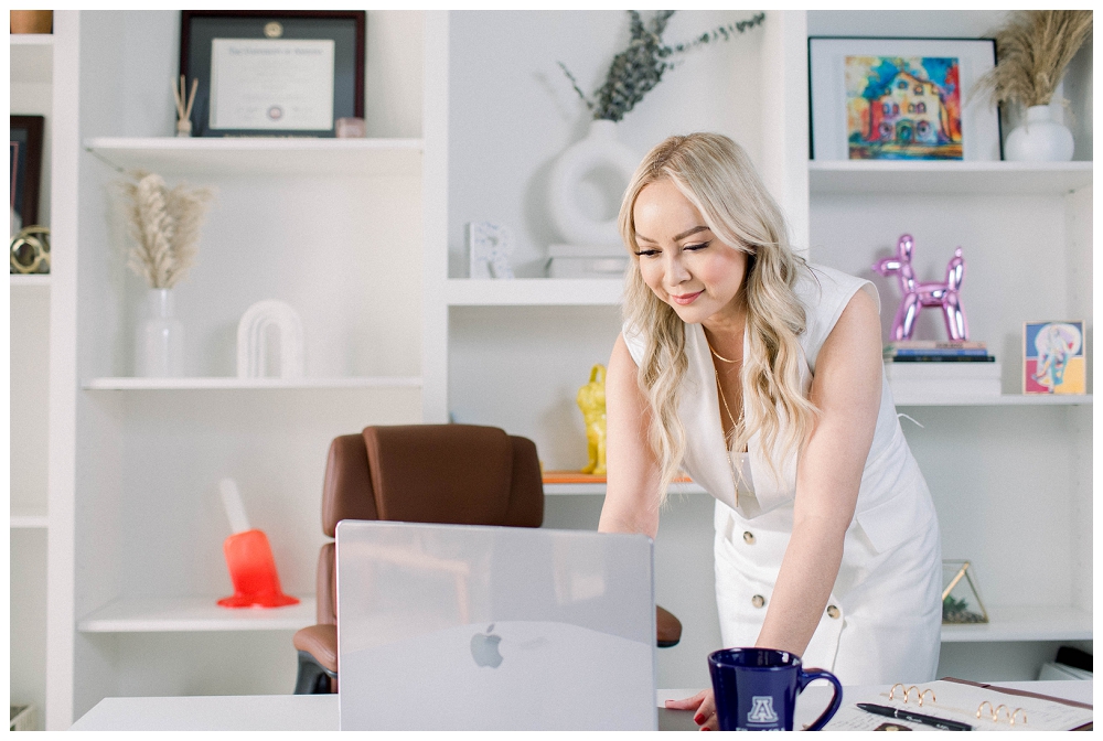 A woman in a beige business outfit leans over a desk and types on a laptop. 
