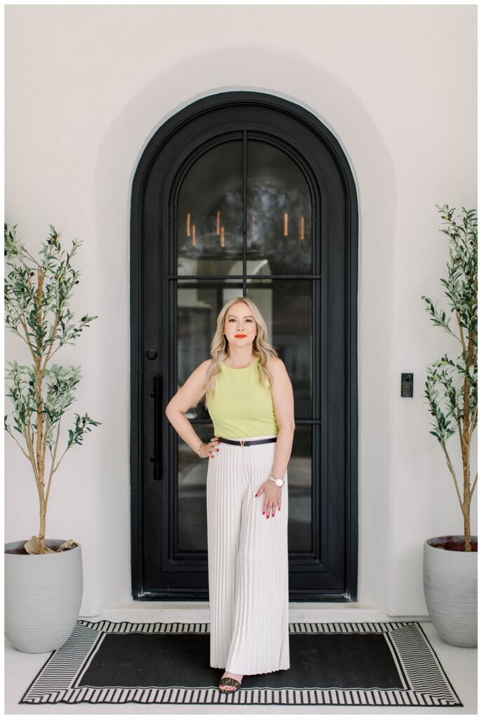 An image of a woman in a green top and white pants, she stands in front of a large black front door. There are two potted trees on either side of her.