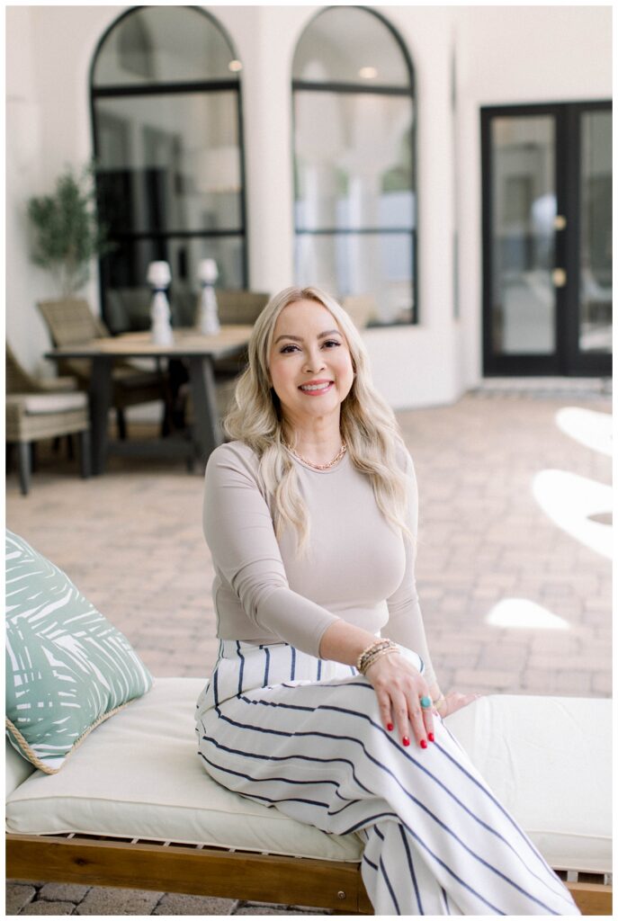 A woman in a beige top and stripped pants sits in an outdoor recliner. She is smiling at the camera. 