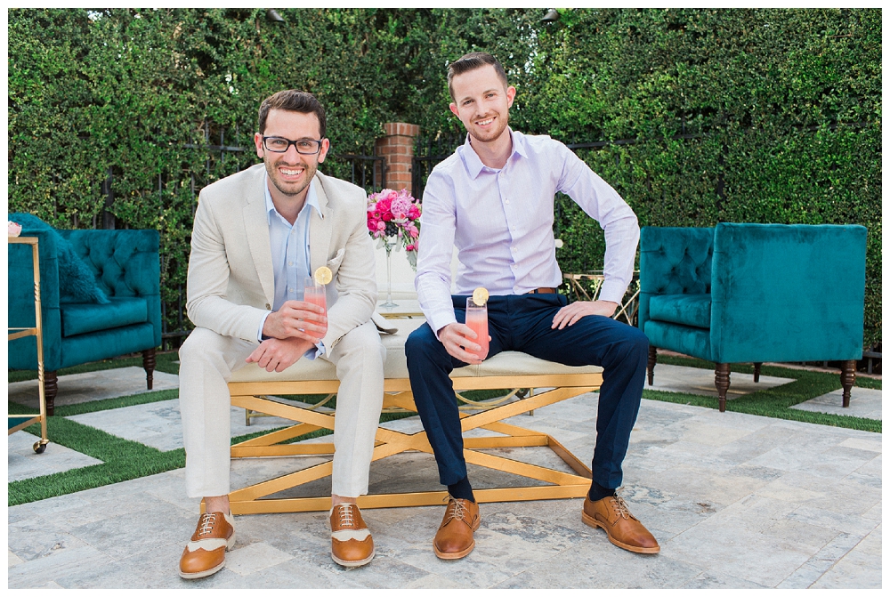 Two well-dressed men sit on a bench holding a pink lemonade. .They smile at the camera. 