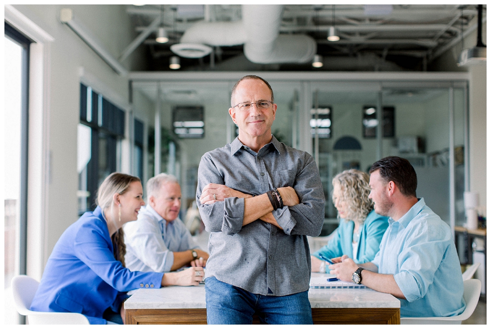 A man in a grey shirt stands in front of a conference table. A team of people sit at the table in the background. 