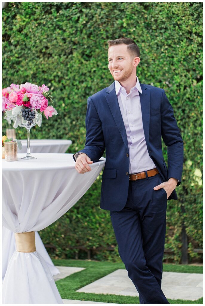 a well dressed man in a navy suit leans slightly on a cocktail table with a floral arrangement on the table.