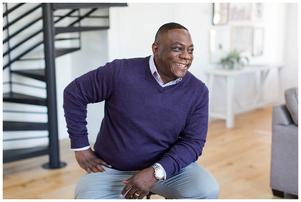 A man in a purple cardigan sits on a stool in a living room and smiles off camera.