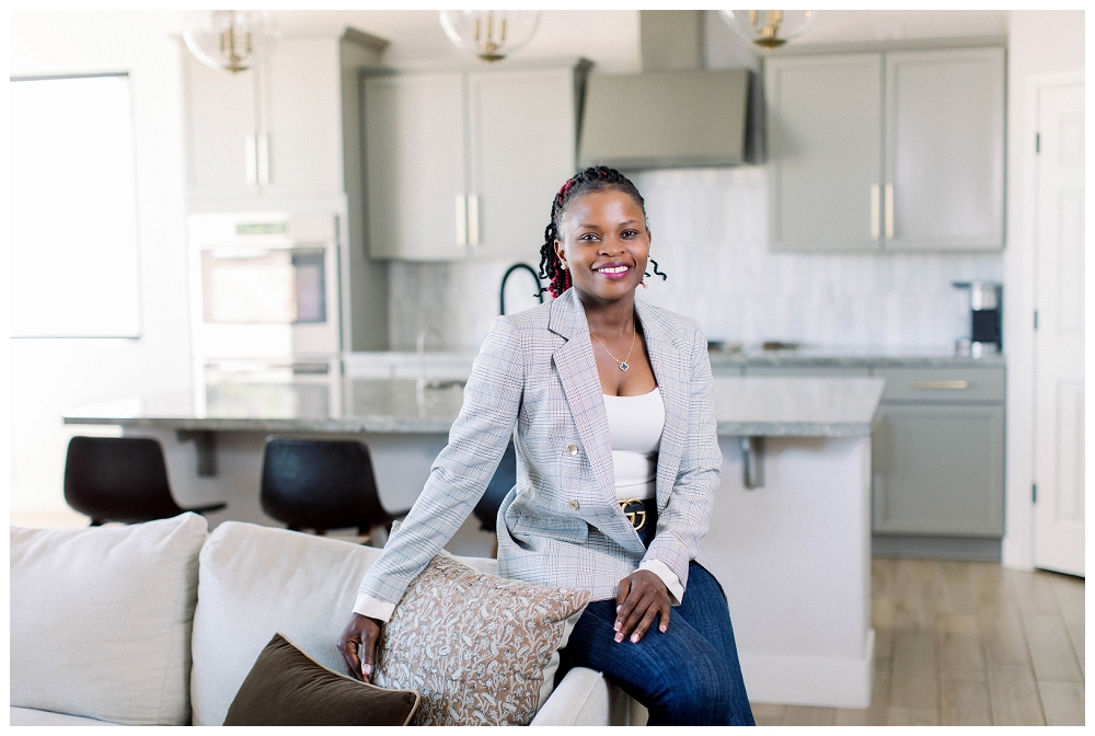 Arizona Brand Photography A woman in a grey blazer and jeans sits on the arm of a sofa and smiles at the camera. There is a kitchen behind her. 