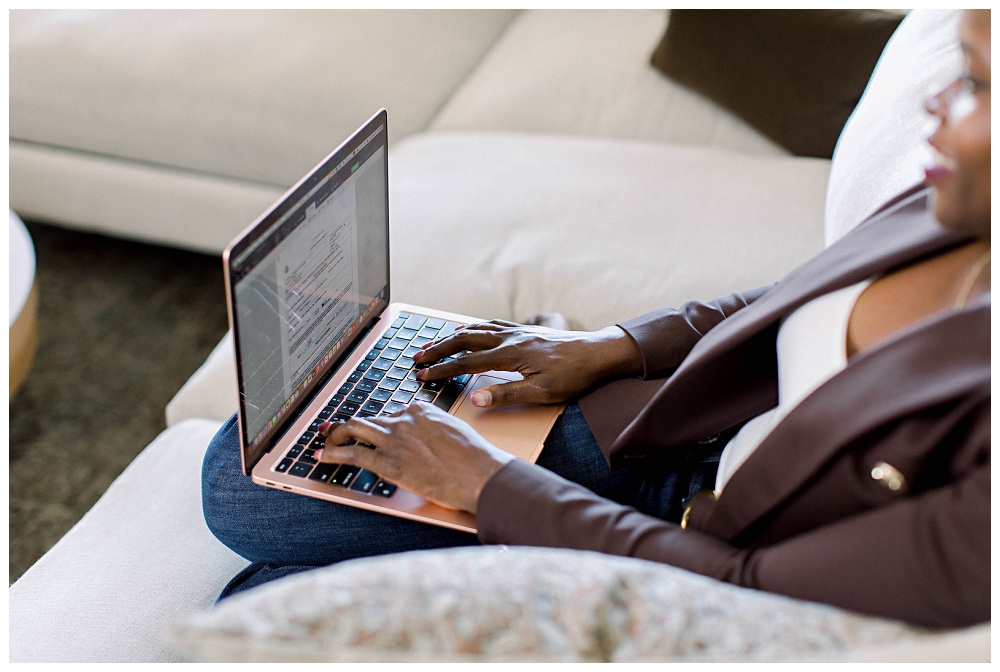 A womain in a brown suit is sitting ona  sofa with a laptop open in her lap. We can see her hands on the keyboard. 