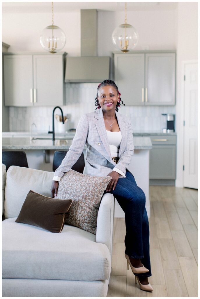 A womain in a grey blazer sits on the arm of a sofa and smiles at the camera. 