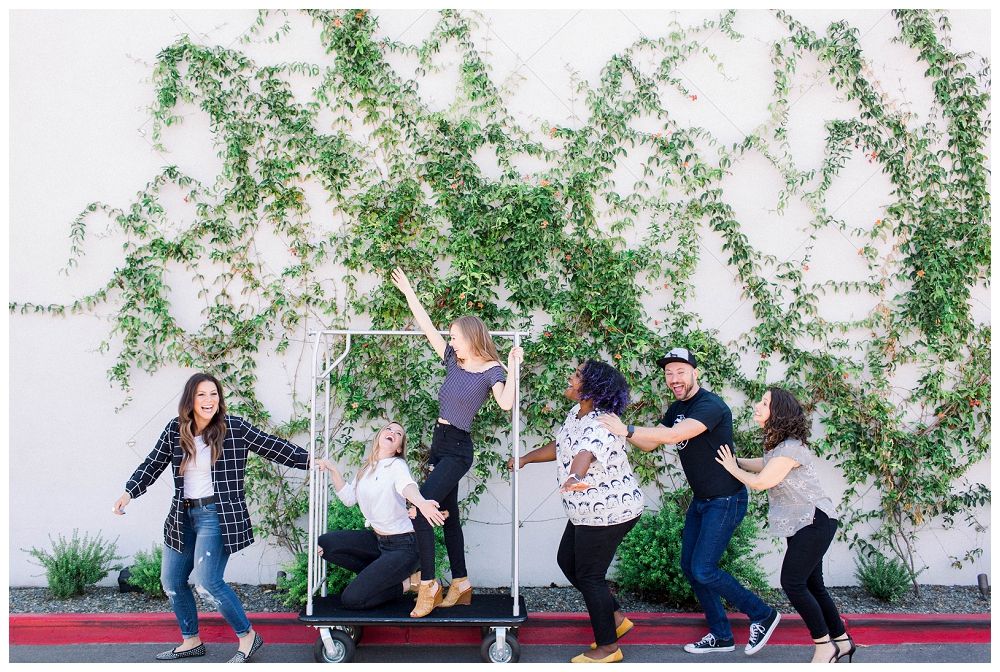 a group of six people are running across the frame of the photo pushing a hotel luggage cart. High energy, fun business team portrait. 