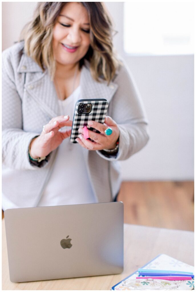 A woman scrolls on a phone standing in front of a laptop