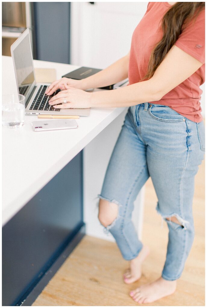 Branding Portrait a woman types on a laptop while standing at a kitchen counter. 