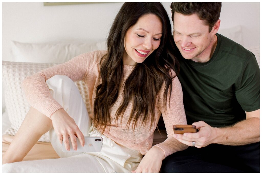 Arizona Brand Photographer couple sits in bed, looking at the husbands cell phone and smiling