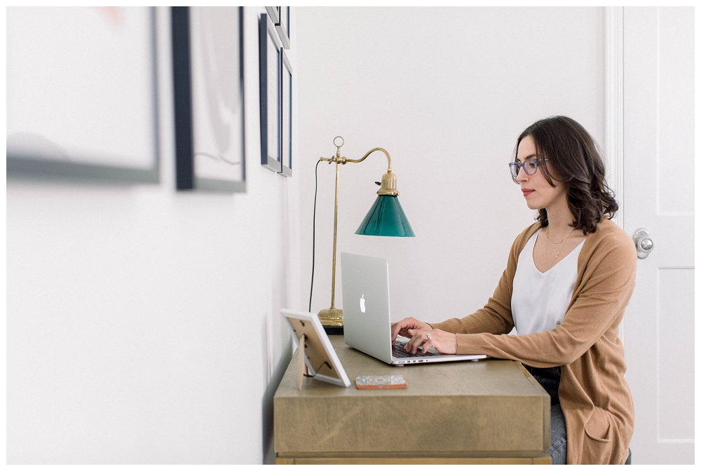 A woman wearing glasses and a beige sweater sits at a desk typing on a laptop.