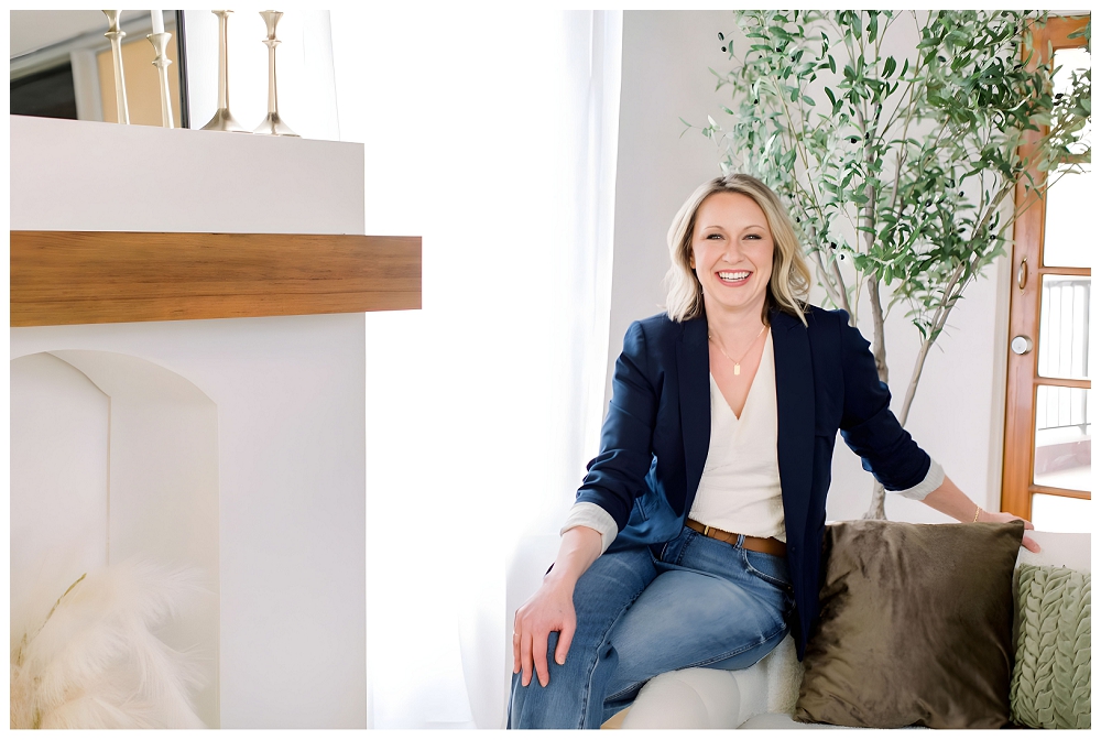 A woman in a navy blazer and jeans sits on the arm of a sofa and smiles at the camera.